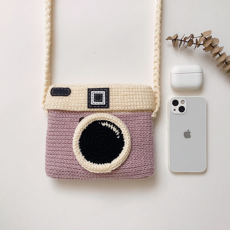 May include: A pink and cream crocheted camera bag with a black lens and a strap.