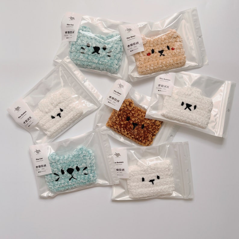 K&ouml;nnte beinhalten: Eine Sammlung handgefertigter H&auml;keltaschen in Tierform in durchsichtiger Plastikverpackung. Die Taschen sind in verschiedenen Farben erh&auml;ltlich, darunter Blau, Beige, Braun und Wei&szlig;, mit gestickten Gesichtern. Jede Tasche ist mit dem Namen des Tieres beschriftet.