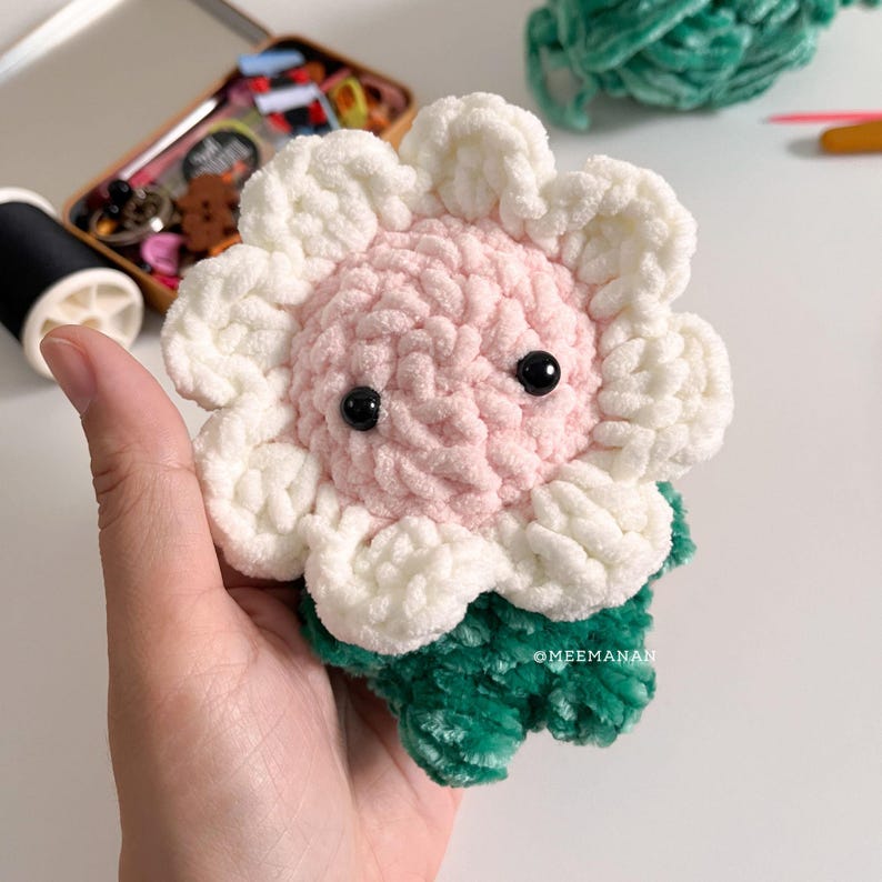 Pu&ograve; includere: Un fiore all'uncinetto fatto a mano con un centro rosa, petali bianchi e foglie verdi. Il fiore ha due occhi di perline nere. Il progetto artigianale &egrave; tenuto in mano. L'immagine include una bobina di filo nero e una piccola scatola di legno con forniture artigianali.