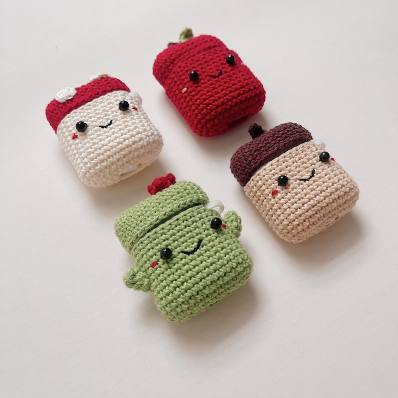 May include: Four crocheted AirPod cases in different shapes and colours: a red apple, a white mushroom, a brown acorn, and a green cactus. Each case has a cute, embroidered face.