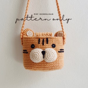May include: A crocheted tiger-shaped bag with a brown strap. The bag has a white face with black eyes and a black nose. The text "PDF DOWNLOAD pattern only" is written above the bag.