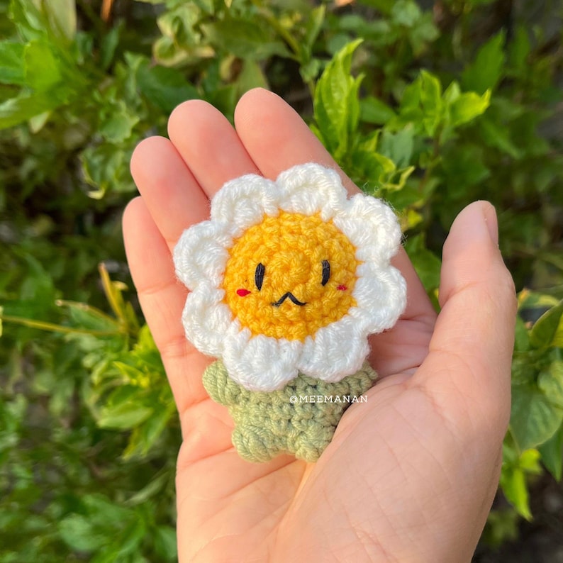 Pu&ograve; includere: Una spilla a forma di fiore all'uncinetto fatta a mano con una faccia sorridente. Il fiore ha petali bianchi, un centro giallo e uno stelo verde. La spilla &egrave; tenuta in mano su uno sfondo di foglie verdi.
