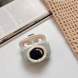 May include: A light green crocheted AirPod case with a black and cream-colored monster mouth design. The case is open, revealing white AirPod earbuds inside. The case is on a white surface with a magazine and crocheted fabric in the background.