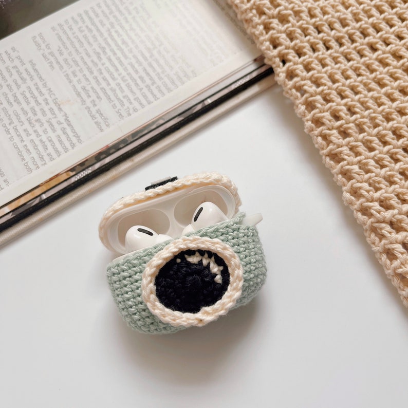 May include: A light green crocheted AirPod case with a black and cream-colored monster mouth design. The case is open, revealing white AirPod earbuds inside. The case is on a white surface with a magazine and crocheted fabric in the background.