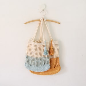 Peut inclure: Deux sacs cabas en crochet suspendus &agrave; un cintre en bois. Le sac du haut est blanc avec un fond bleu clair et un pompon bleu clair. Le sac du bas est jaune uni avec un pompon jaune.