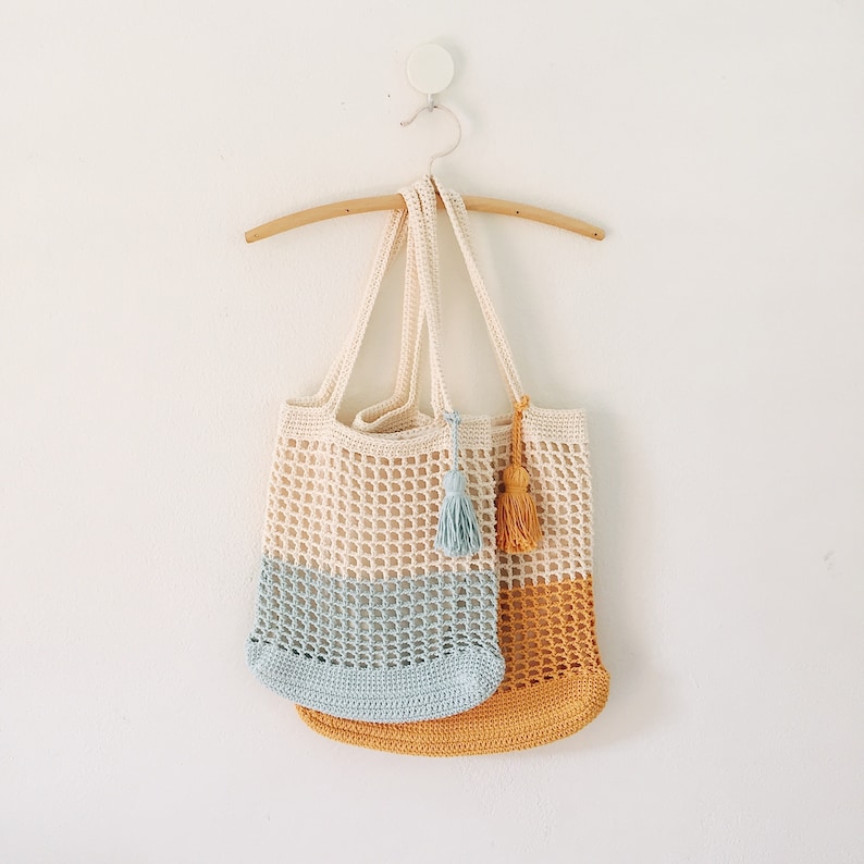 Peut inclure: Deux sacs cabas en crochet suspendus &agrave; un cintre en bois. Le sac du haut est blanc avec un fond bleu clair et un pompon bleu clair. Le sac du bas est jaune uni avec un pompon jaune.