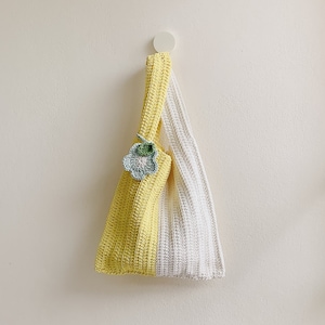 Peut inclure: Un sac cabas en crochet jaune et blanc avec une petite fleur en crochet comme embellissement. Le sac est suspendu &agrave; un crochet blanc sur un mur blanc.