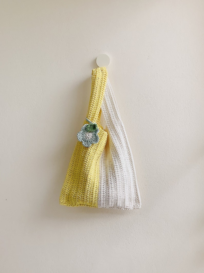Peut inclure: Un sac cabas en crochet jaune et blanc avec une petite fleur en crochet comme embellissement. Le sac est suspendu &agrave; un crochet blanc sur un mur blanc.