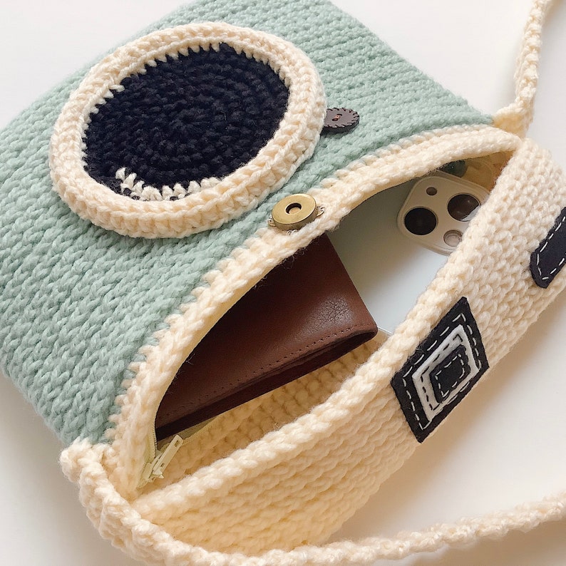 May include: A crocheted camera-shaped bag with a light blue and cream color scheme. The bag has a brown leather wallet and a white phone inside. The bag has a long strap and a black and white crocheted design on the front.