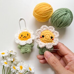 Pu&ograve; includere: Due portachiavi a margherita all'uncinetto fatti a mano con facce sorridenti, petali bianchi e steli verdi. Un gomitolo di lana giallo e verde &egrave; sullo sfondo. I portachiavi sono alti circa 7.6 cm.