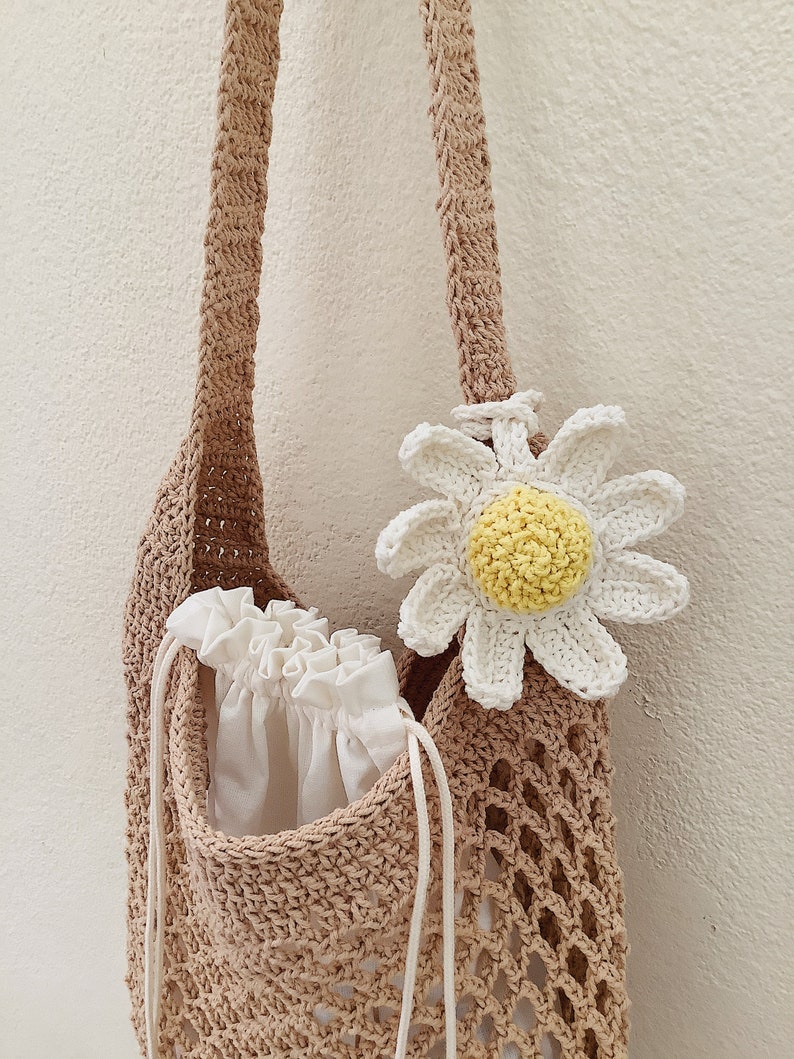 Peut inclure: Un sac cabas en crochet marron avec une fleur de marguerite en crochet blanc attach&eacute;e &agrave; la bandouli&egrave;re. Le sac est doubl&eacute; d'une doublure blanche &agrave; cordon et d'une bandouli&egrave;re en crochet blanc.