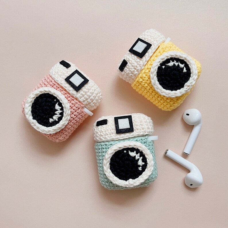 May include: Three crochet camera cases in pastel colors: pink, yellow, and blue. Each case has a white top with a black square detail and a large black and white lens. White wireless earbuds are next to the cases.