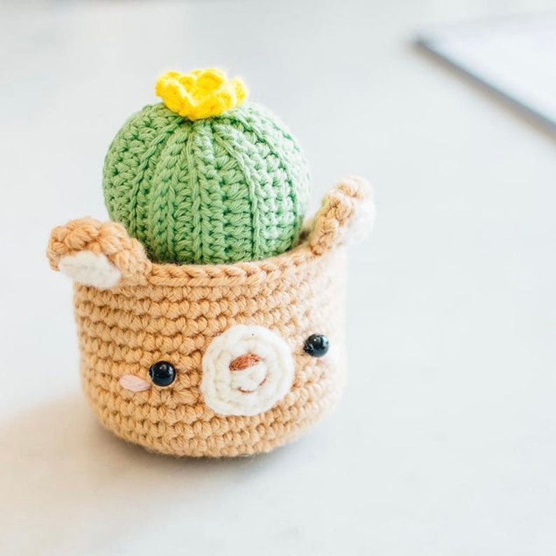 Puede incluir: Una planta de cactus de crochet en una maceta de crochet marr&oacute;n con una linda cara de oso. El cactus es verde con una flor amarilla en la parte superior.
