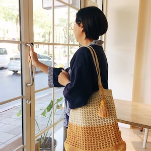 Peut inclure: Une femme portant une chemise bleue et des lunettes tient un sac fourre-tout en crochet avec un motif jaune et blanc. Le sac a un pompon sur le c&ocirc;t&eacute;. Le panneau au-dessus de la porte indique "Craft Melt."