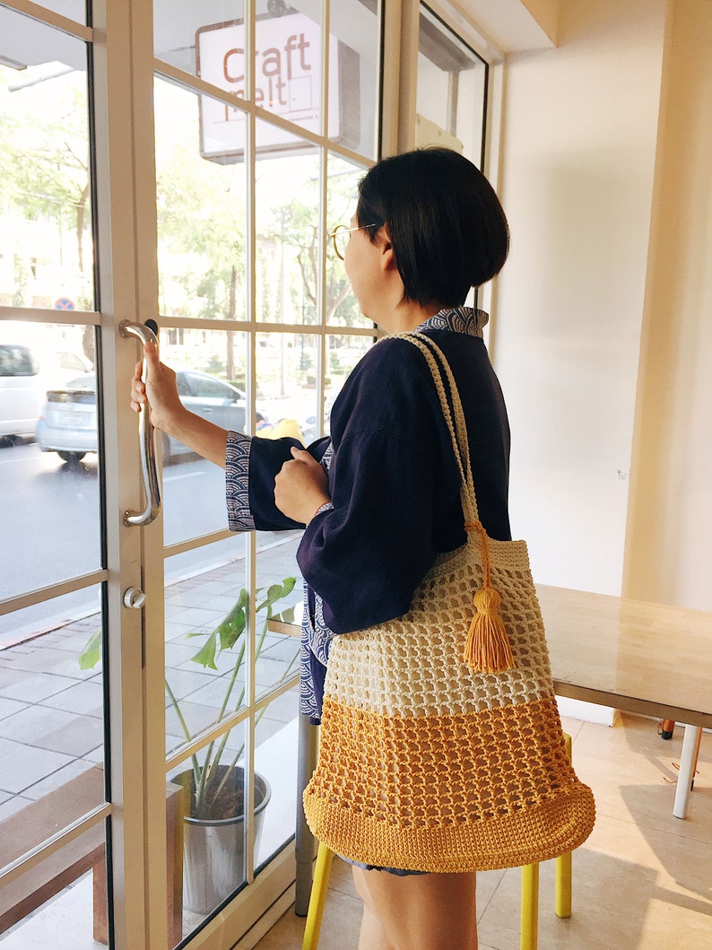 Peut inclure: Une femme portant une chemise bleue et des lunettes tient un sac fourre-tout en crochet avec un motif jaune et blanc. Le sac a un pompon sur le c&ocirc;t&eacute;. Le panneau au-dessus de la porte indique "Craft Melt."