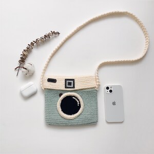 May include: A light green and cream crocheted camera bag with a long cream strap. The bag has a black lens and a brown strap detail.