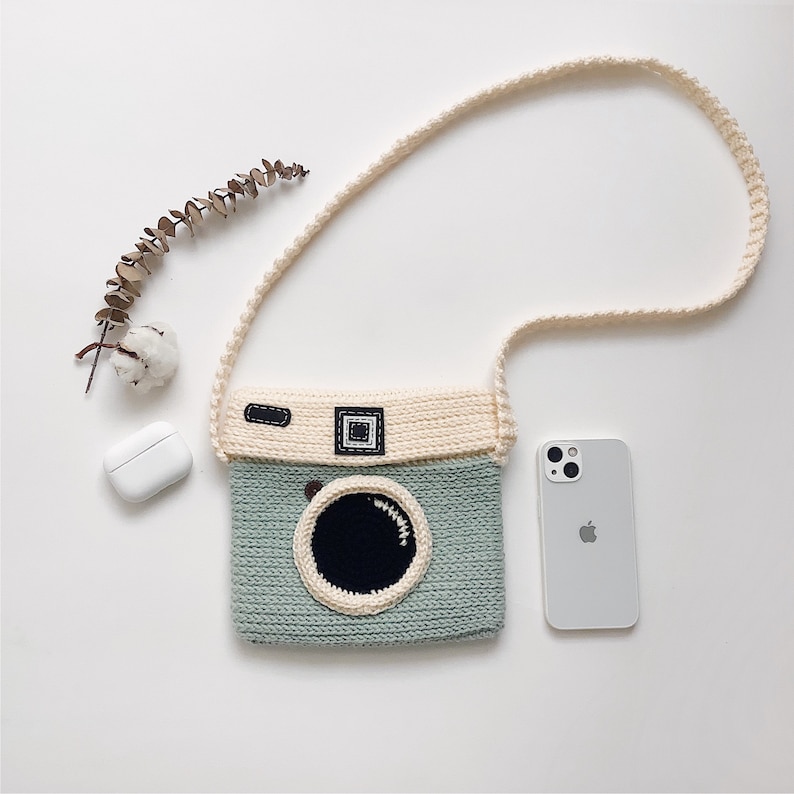 May include: A light green and cream crocheted camera bag with a long cream strap. The bag has a black lens and a brown strap detail.