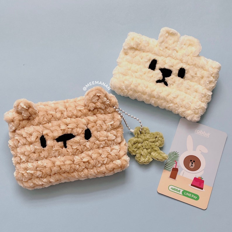 K&ouml;nnte beinhalten: Zwei handgefertigte, pl&uuml;schige, b&auml;renf&ouml;rmige H&uuml;llen in Beige und Creme mit schwarzen, gestickten Gesichtsmerkmalen. Ein gr&uuml;ner Kleeblatt-Anh&auml;nger und eine Rabbit LINE Pay-Karte sind ebenfalls vorhanden. Die H&uuml;llen sind wahrscheinlich f&uuml;r kleine elektronische Ger&auml;te.
