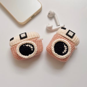 May include: Two pink and cream-colored crochet camera-shaped AirPod cases. Each case features a black lens, a white border, and a small black square on top. White earbuds and a smartphone are also visible.