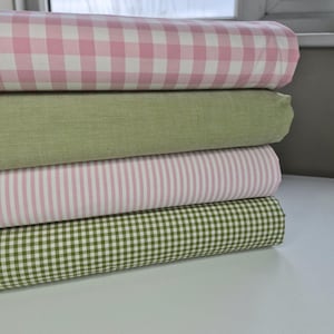 Puede incluir: Una pila de rollos de tela en varios patrones y colores. El rollo superior presenta un estampado de cuadros rosa y blanco, seguido de una tela verde lisa, una tela a rayas rosa y blanca y un estampado de cuadros verde y blanco en la parte inferior.