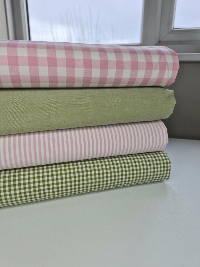 Puede incluir: Una pila de rollos de tela en varios patrones y colores. El rollo superior presenta un estampado de cuadros rosa y blanco, seguido de una tela verde lisa, una tela a rayas rosa y blanca y un estampado de cuadros verde y blanco en la parte inferior.