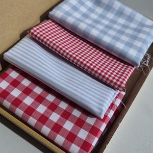 May include: A cardboard box contains four folded fabric pieces. The fabrics feature red and white gingham, light blue and white gingham, and light blue and white striped patterns. The fabrics are neatly arranged inside the box.