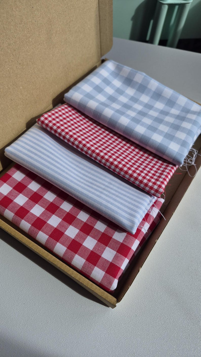 May include: A cardboard box contains four folded fabric pieces. The fabrics feature red and white gingham, light blue and white gingham, and light blue and white striped patterns. The fabrics are neatly arranged inside the box.