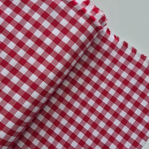 May include: Red and white checkered fabric, folded and displayed. The fabric has a classic gingham pattern, with a frayed edge visible. The material is likely cotton or a similar woven textile, suitable for various sewing projects.