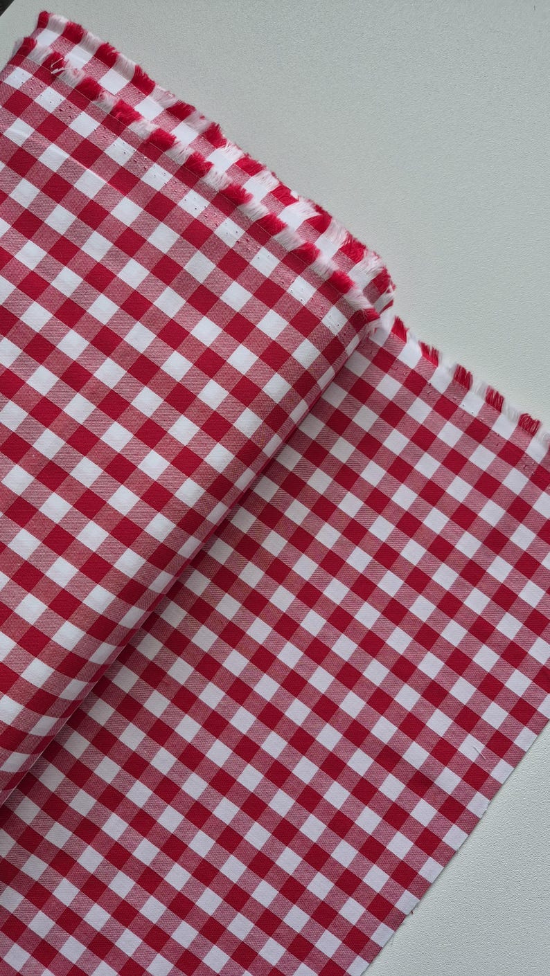 May include: Red and white checkered fabric, folded and displayed. The fabric has a classic gingham pattern, with a frayed edge visible. The material is likely cotton or a similar woven textile, suitable for various sewing projects.