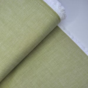 Puede incluir: Un rollo de tela verde claro con una superficie texturizada. La tela est&aacute; doblada, revelando un borde blanco con flecos. El material parece ser lino o un textil tejido similar, adecuado para proyectos de ropa o decoraci&oacute;n del hogar.
