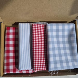 May include: A cardboard box containing four folded fabric squares. The fabrics feature red and white gingham, blue and white stripes, red and white gingham, and blue and white gingham patterns. The fabrics are likely cotton or a similar material.