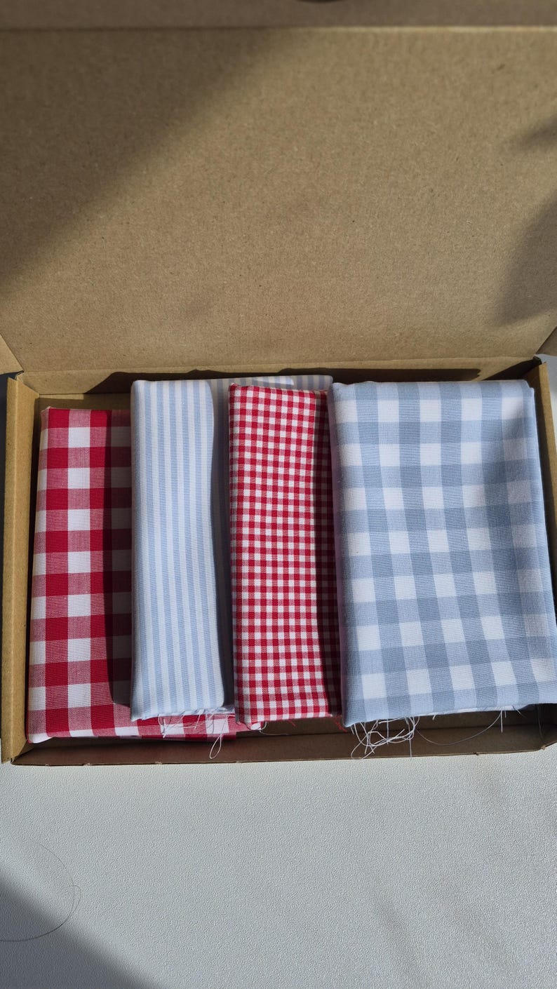 May include: A cardboard box containing four folded fabric squares. The fabrics feature red and white gingham, blue and white stripes, red and white gingham, and blue and white gingham patterns. The fabrics are likely cotton or a similar material.