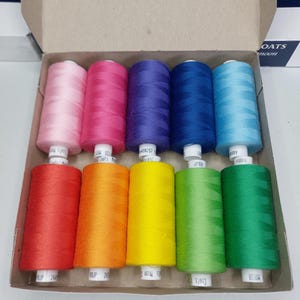 May include: A selection of Coats thread spools in a cardboard box, showcasing a spectrum of colours. The spools are in shades of pink, fuchsia, purple, blue, teal, red, orange, yellow, green, and dark green. Each spool has a white label.