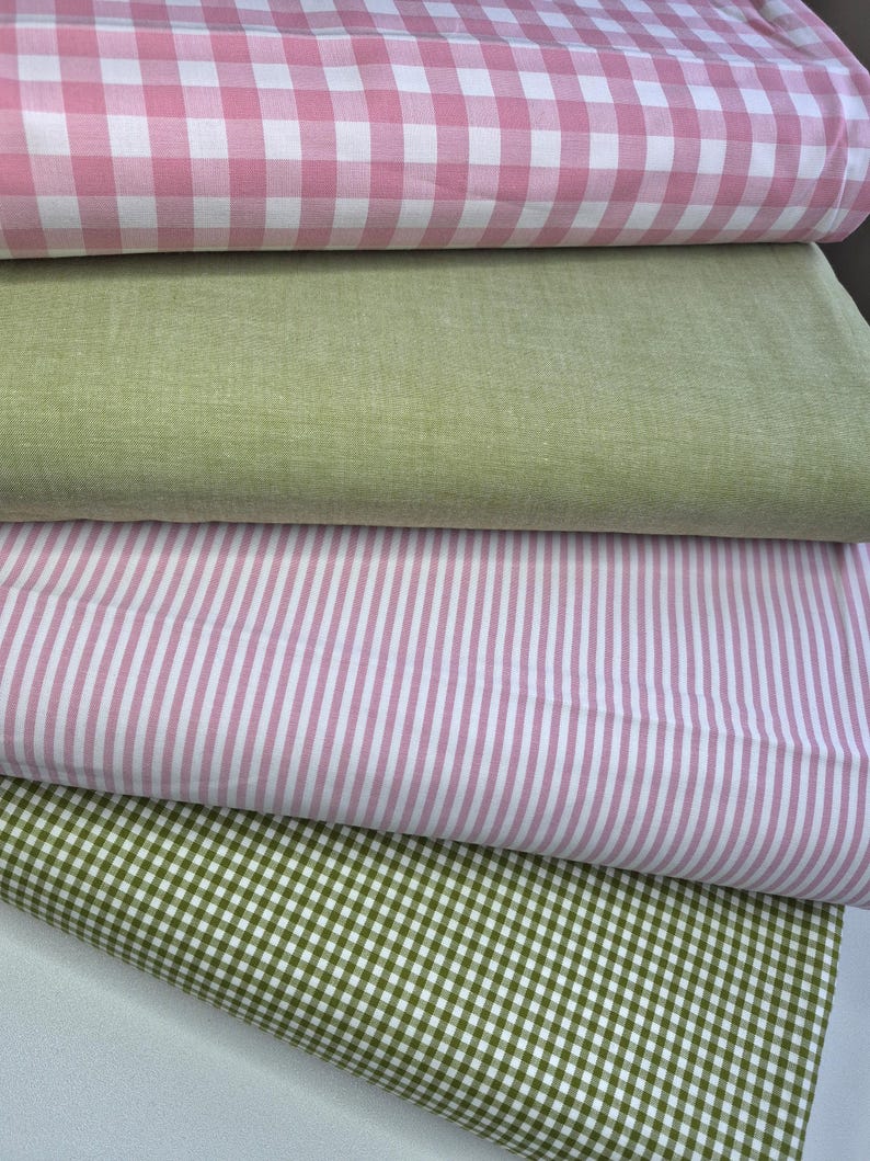 Puede incluir: Una pila de muestras de tela en varios patrones y colores. La capa superior presenta un estampado de cuadros rosa y blanco, seguida de una tela verde claro lisa. Debajo hay una tela a rayas rosa y blanca y una tela a cuadros verde y blanca.