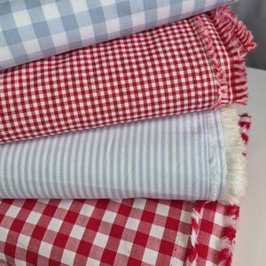May include: A stack of fabric samples in various patterns. The top layer is light blue and white gingham, followed by red and white gingham, light blue and white stripes, and red and white gingham. The edges of the red fabrics have a fringed detail.