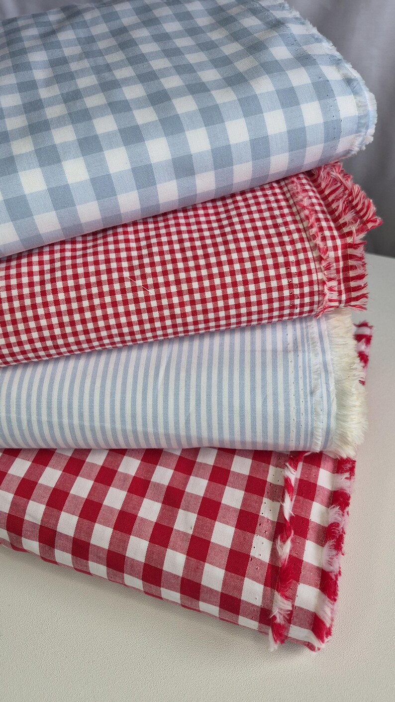 May include: A stack of fabric samples in various patterns. The top layer is light blue and white gingham, followed by red and white gingham, light blue and white stripes, and red and white gingham. The edges of the red fabrics have a fringed detail.