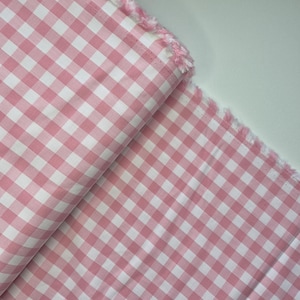 Puede incluir: Tela de cuadros vichy rosa y blanco con un borde deshilachado. La tela est&aacute; doblada, mostrando el patr&oacute;n de cuadrados espaciados uniformemente. El color es un rosa suave, y la tela parece ser de algod&oacute;n o un material similar.