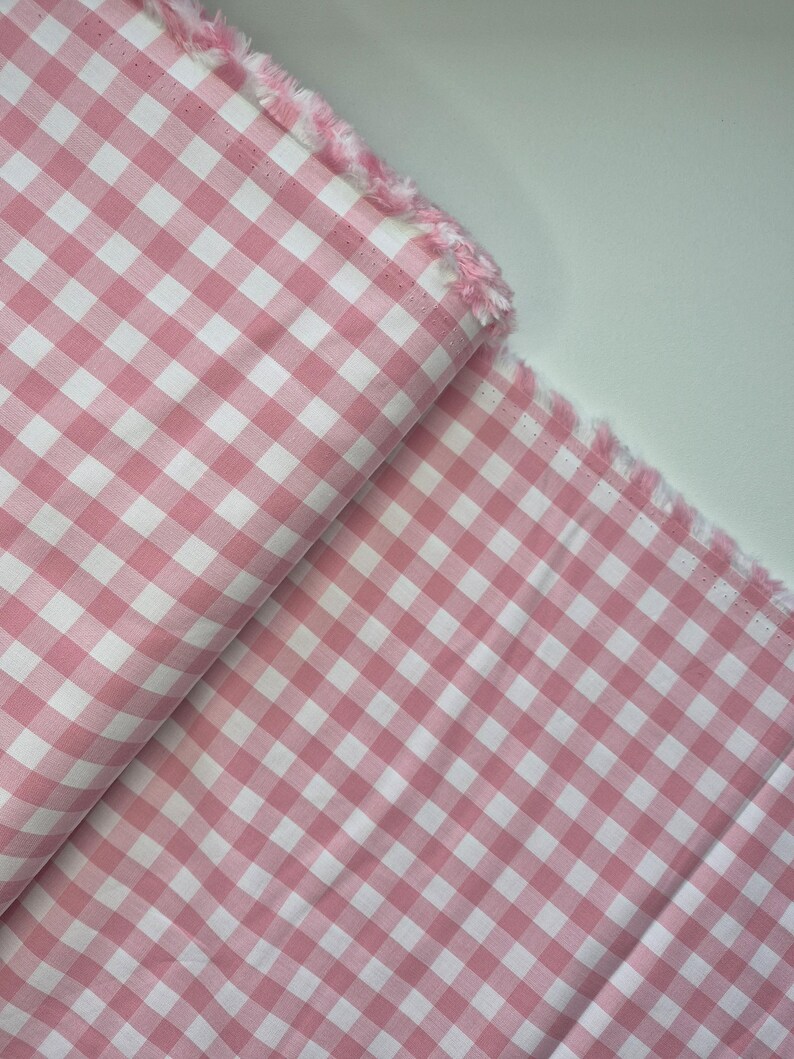 Puede incluir: Tela de cuadros vichy rosa y blanco con un borde deshilachado. La tela est&aacute; doblada, mostrando el patr&oacute;n de cuadrados espaciados uniformemente. El color es un rosa suave, y la tela parece ser de algod&oacute;n o un material similar.