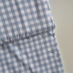 May include: Close-up of a light blue and white gingham fabric. The fabric is folded, revealing a layered effect. The edges are unfinished, with visible stitching and frayed threads. The pattern is a classic checkered design.