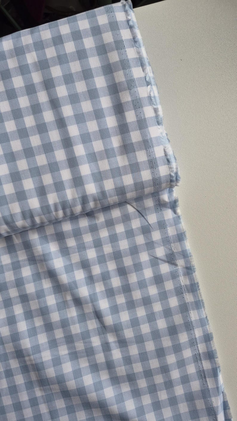 May include: Close-up of a light blue and white gingham fabric. The fabric is folded, revealing a layered effect. The edges are unfinished, with visible stitching and frayed threads. The pattern is a classic checkered design.