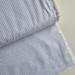 May include: Close-up of a folded fabric with a blue and white striped pattern. The fabric has a raw, unfinished edge with visible threads. The stripes run vertically across the material, creating a classic design.