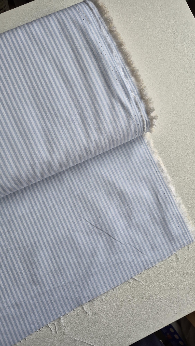 May include: Close-up of a folded fabric with a blue and white striped pattern. The fabric has a raw, unfinished edge with visible threads. The stripes run vertically across the material, creating a classic design.
