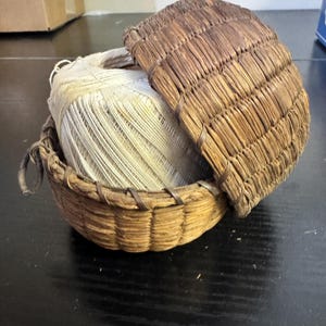 Op de afbeelding: Een kleine, ronde, geweven mand met een deksel, gevuld met een grote spoel wit garen. De mand en het deksel zijn gemaakt van natuurlijk gekleurd riet en zijn vastgezet met een donkerbruine binding.