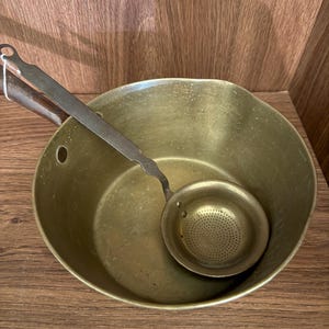Op de afbeelding: Een vintage messing kookpot met een bijpassende zeeflepel. De pot heeft een afgeronde vorm met een handvat en de zeeflepel heeft een geperforeerde kom. De items zijn gemaakt van messing en hebben een warme, antieke uitstraling.