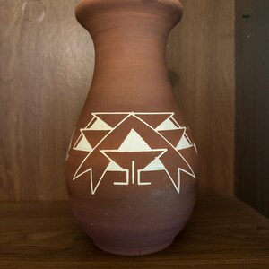 May include: A terracotta vase with a wide base and a narrow neck. The vase has a white geometric pattern around the middle. The vase is set against a wooden background.