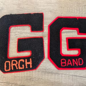 May include: Two black varsity letter patches shaped like the letter "G". One patch has "ORCH" in orange lettering, and the other has "BAND" in orange lettering. Both have a red border.