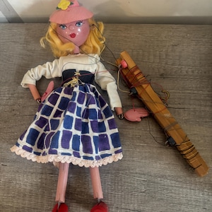 May include: A vintage marionette doll with blonde hair, a pink hat, and a white top. The doll has a blue and purple checkered skirt, pink legs, and red shoes. The wooden control is next to the doll.
