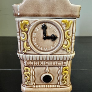 Puede incluir: Un tarro de galletas de cerámica con forma de torre del reloj. El tarro es de color beige con detalles amarillos y marrones. La esfera del reloj marca las 9 en punto. Las palabras "COOKIE TIME" están impresas en la parte delantera.