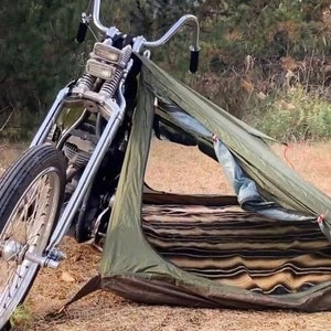 May include: A classic motorcycle with a silver frame and black tyres stands beside a small olive green tent. The tent is open, showing a striped interior. The scene is set outdoors with dry grass and foliage.