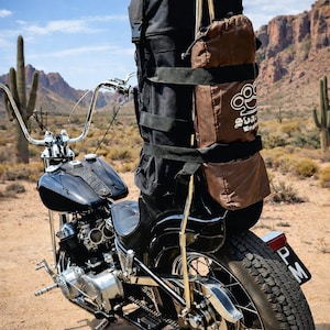 Puede incluir: Una motocicleta negra con una gran bolsa negra y bolsas marrones adjuntas en la parte trasera. La motocicleta tiene manillar cromado y un depósito de combustible negro. El fondo presenta un paisaje desértico con cactus y montañas.